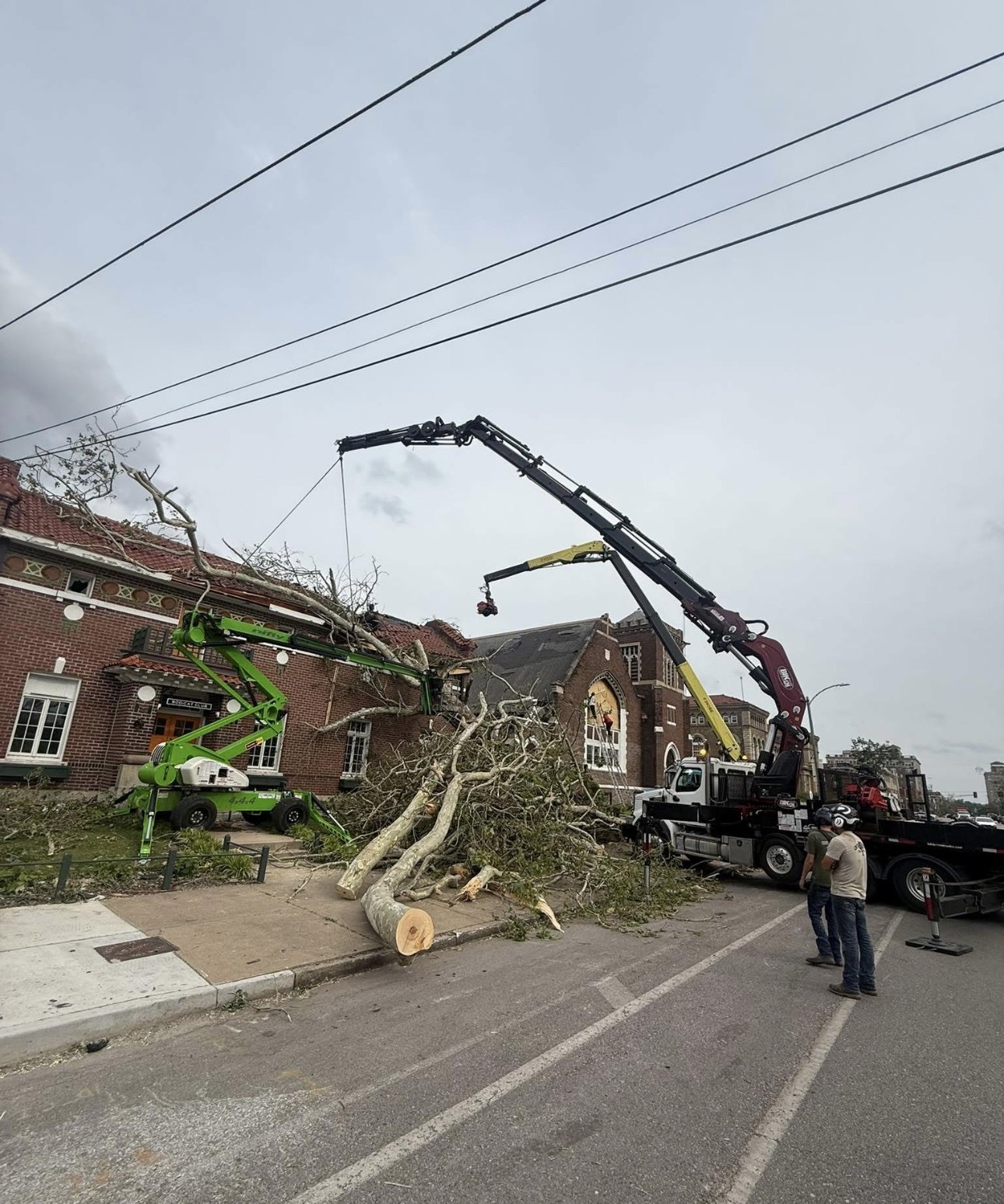 Grapple crane and green boom lift clearing a fallen tree from a brick building as crew watch from the street