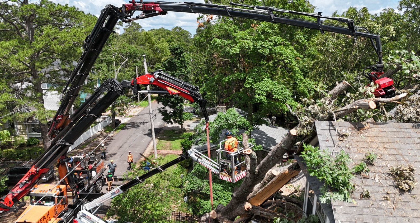 Aerial view of two grapple-saw cranes and a bucket lift removing a large tree from a house roof